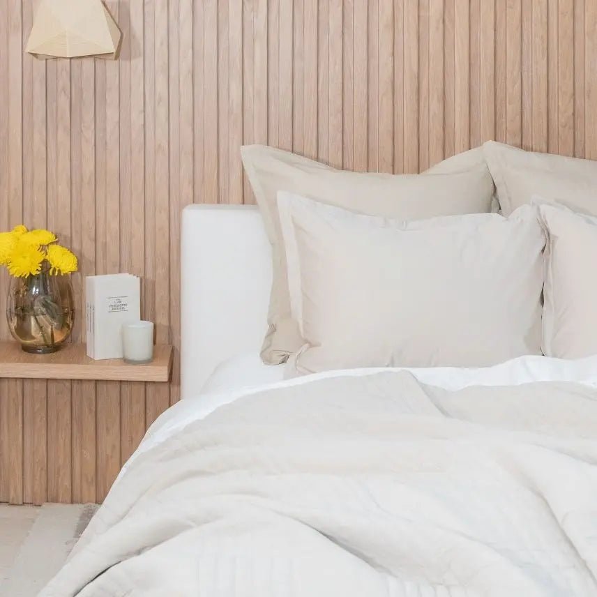 Neatly made bed with beige bedding and beige pillows against a wooden wall.