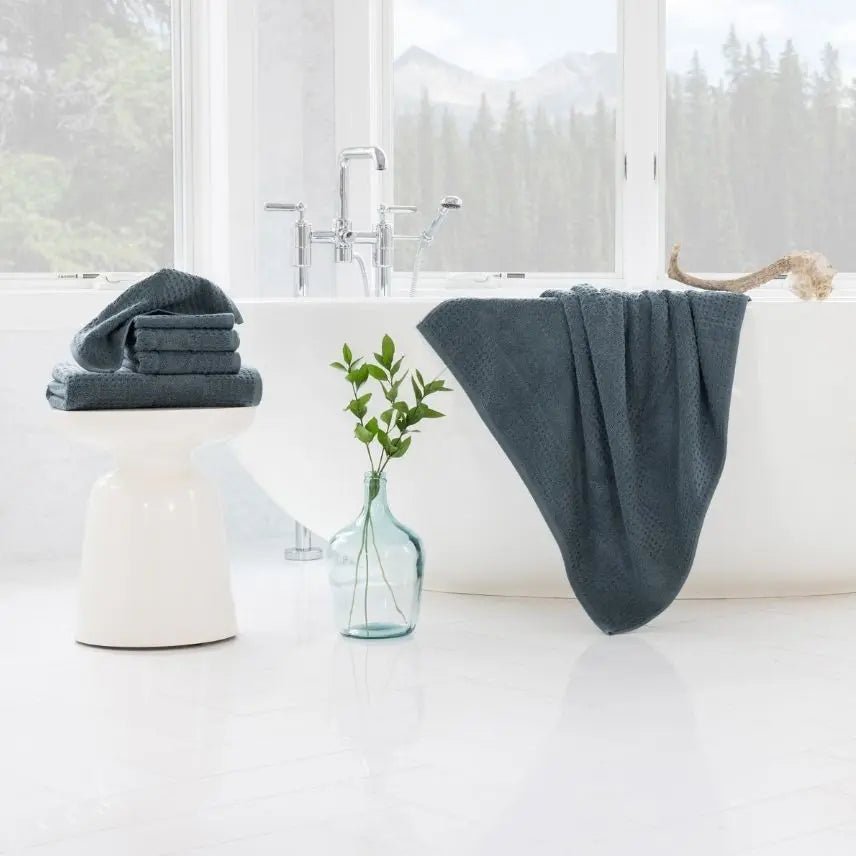 Bathroom scene with blue teal towels on a white bathtub and stool, and a vase with greenery.